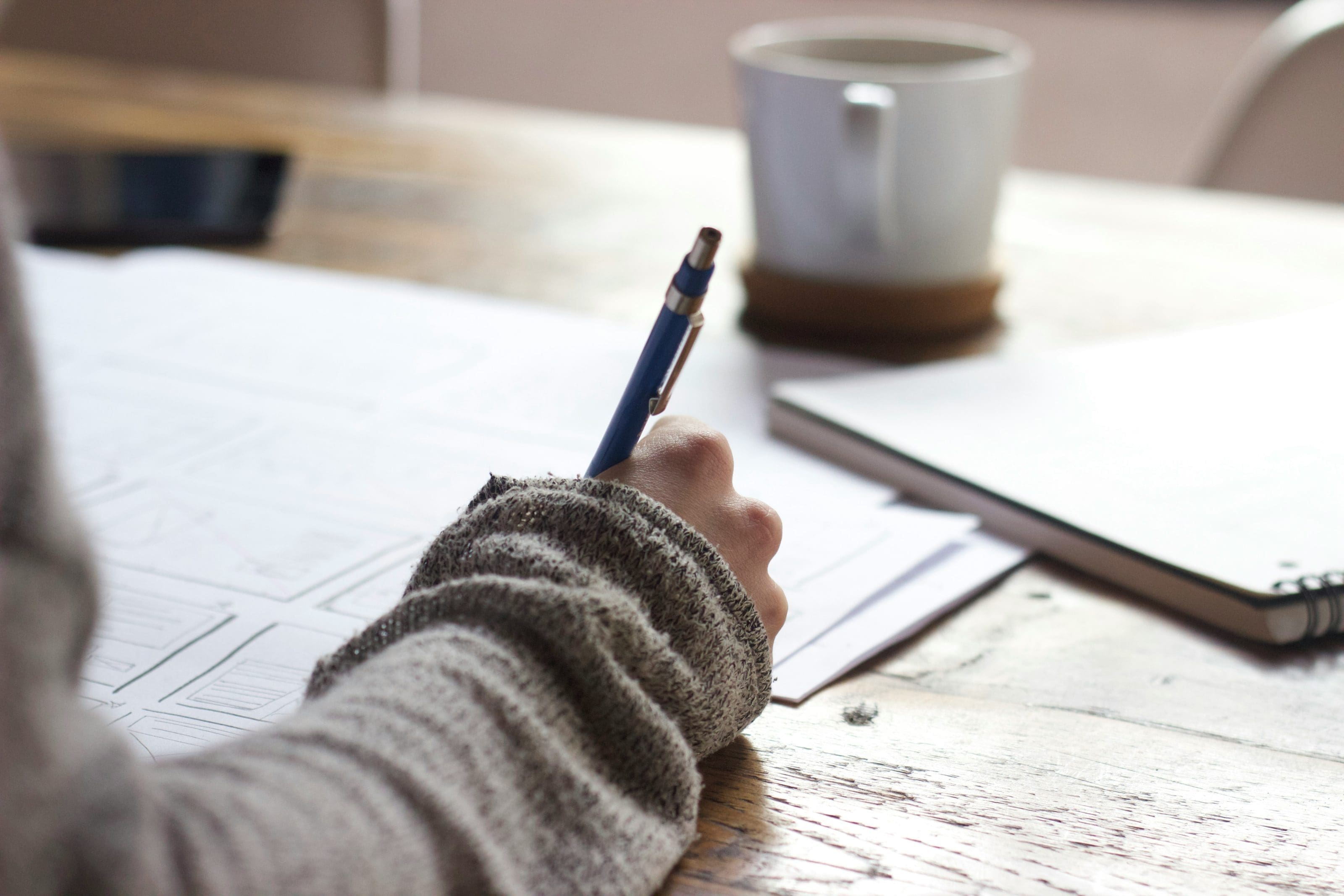 a person writing onto paper with a pen. a cup of coffee in the background